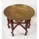 A LARGE INDO - CHINESE HAND CHASED CIRCULAR BRASS TRAY, sat upon a six-legged wooden folding