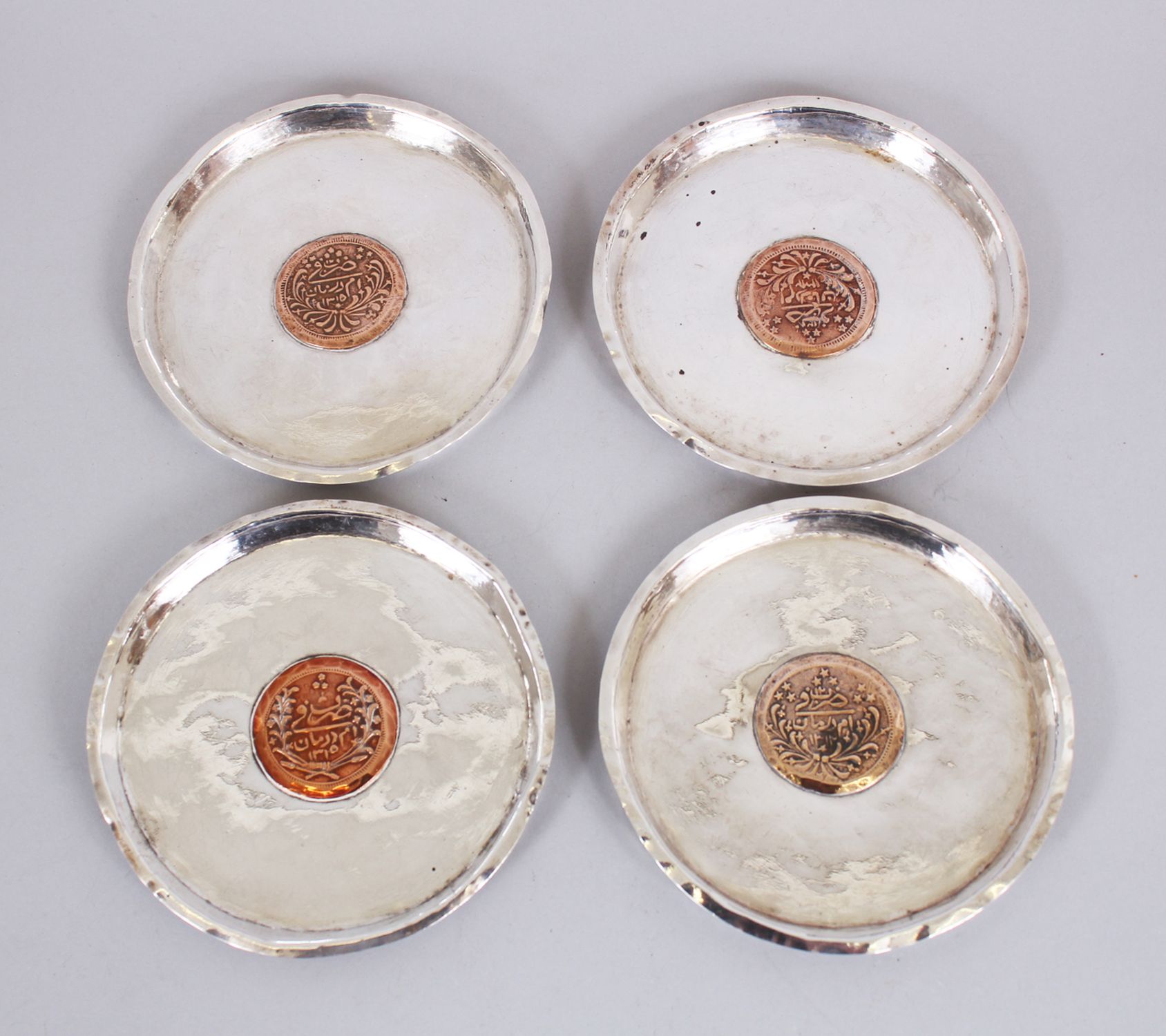 A SET OF FOUR ISLAMIC SILVER CIRCULAR DISHES inserted with coins, 11cm diameter.