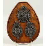 THREE CAST METAL ARMORIAL CRESTS, on an oak plaque. 24cms high.
