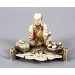 A JAPANESE MEIJI PERIOD CARVED IVORY OKIMONO OF A FRUIT SELLER, the man seated upon a carved base,