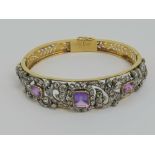 A yellow metal hinged bangle having three graduated amethyst and pierced scrolling frond design