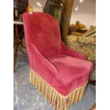 A LATE VICTORIAN WINE RED CORDUROY UPHOLSTERED LOW ARMCHAIR WITH TASSELLED APRON HANGING OVER THE
