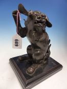 A BRONZE FIGURE OF A BEGGING BUDDHIST LION LOOKING OVER HIS LEFT SHOULDER AND MOUNTED ON AN EBONISED