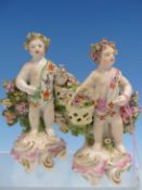 A PAIR OF DERBY PORCELAIN PUTTI CARRYING BASKETS OF FLOWERS AND BACKED BY BOCAGES. H 13.5cms.