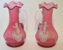 Two cranberry coloured glass ewers or vases with loop handles decorated in Mary Gregory style with