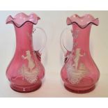 Two cranberry coloured glass ewers or vases with loop handles decorated in Mary Gregory style with