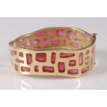 A yellow metal hinged oval bangle, featuring a wavy design with assorted apertures filled with red