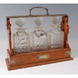 A late Victorian oak and silver plated three bottle tantalus, the square cut decanters and