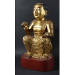 SOUTH EAST ASIAN CARVED AND GILTWOOD FEMALE DEITY, modelled seated with one hand raised, the other