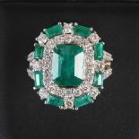 A white metal emerald and diamond cocktail ring, centred with an emerald cut emerald, calculated