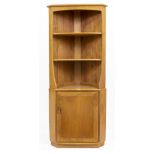 AN ERCOL LIGHT ELM CORNER CABINET with two fixed shelves above a cupboard door below, raised on a