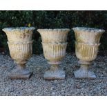 A SET OF THREE RECONSTITUTED STONE GARDEN URNS with flowerhead moulded designs on fluted bases (