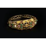 A VARI GEM-SET BANGLE, of hinged oval form, decorated with flowerheads, foliage and scrolls,