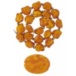 A NECKLACE OF TWENTY TWO IRREGULAR AMBER BEADS WITH SILVERED METAL SPACERS AND A CARVED AMBER BROOCH