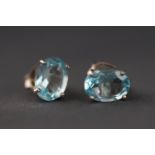 A white metal pair of single stone stud earrings each set with an oval faceted cut blue topaz.