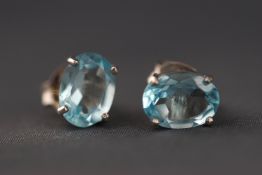 A white metal pair of single stone stud earrings each set with an oval faceted cut blue topaz.