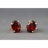 A white metal pair of single stone studs each set with an oval faceted cut garnet.