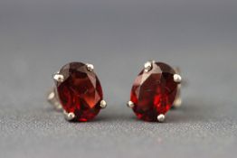A white metal pair of single stone studs each set with an oval faceted cut garnet.
