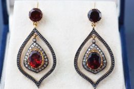 A yellow and white metal pair of drop earrings. Each set with garnets and single cut diamonds.