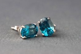 A white metal pair of single stone stud earrings each set with an oval faceted cut blue topaz.