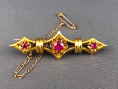 A yellow metal bar brooch set with three graduated round faceted cut rubies.