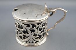 A pierced silver cylindrical lidded mustard pot with strap thumb piece and scroll handle,