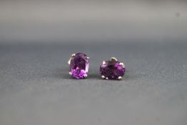 A white metal pair of single stone studs set with an oval faceted cut amethyst