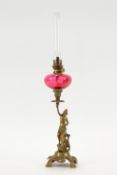 A brass oil lamp in the form of a lady holding aloft a cranberry glass reservoir,