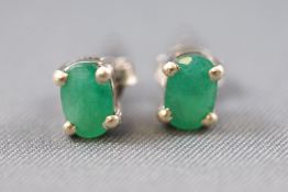 A white metal pair of single stone studs, each set with an oval faceted cut emerald.