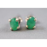 A white metal pair of single stone studs, each set with an oval faceted cut emerald.