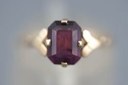 A yellow metal single stone ring set with a rectangular faceted cut amethyst.