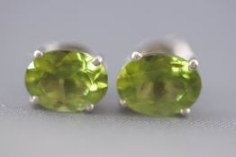 A white metal pair of single stone stud earrings each set with an oval faceted cut peridot.