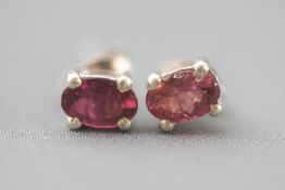 A white metal pair of single stone stud earrings. Each set with an oval faceted cut pink tourmaline.