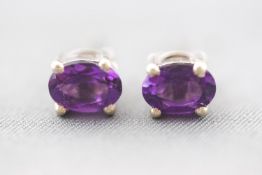 A white metal pair of single stone stud earrings. Each set with an oval faceted cut amethyst.