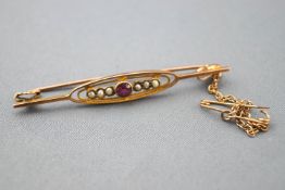 A yellow metal bar brooch set with a central round cut ruby and finished with seed pearls.
