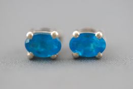 A white metal pair of single stone stud earrings. Each set with an oval faceted cut blue apatite.