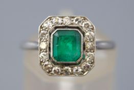 A white metal cluster ring. Principally set with a rectangular cut emerald estimated to weigh 0.