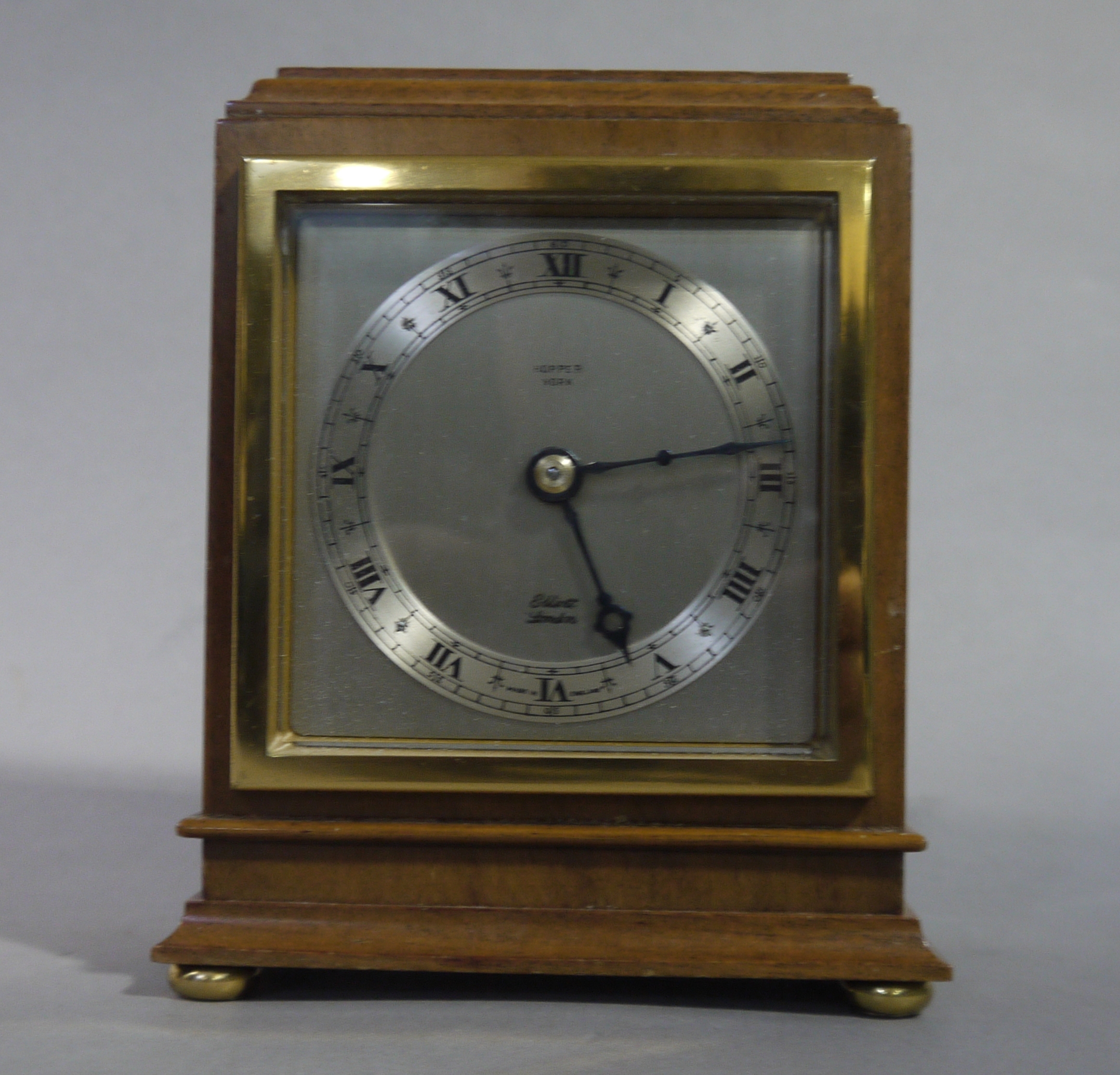 An Elliott walnut mantel clock of square stepped outline with brass bezel and bun feet,