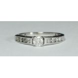 An 18ct white gold solitaire diamond ring four claw-set with a round RBC diamond and channel set