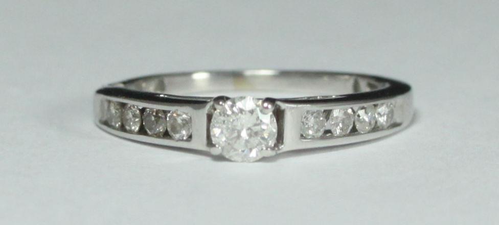 An 18ct white gold solitaire diamond ring four claw-set with a round RBC diamond and channel set