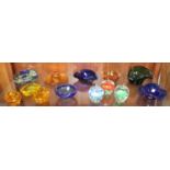 A shelf of coloured glass ashtrays together with three clear glass paperweights modelled as apples