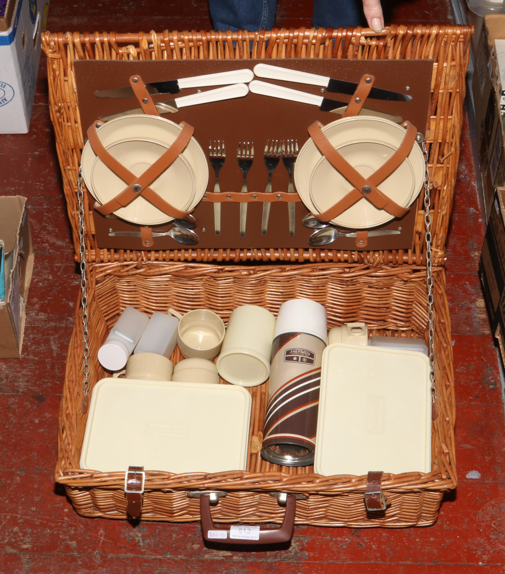 A wicker picnic hamper with contents of stainless steel cutlery and food storage containers.