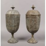 A pair of Indian lidded brass pedestal urns chased with figures and deities.