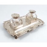 A silver desk stand, of shaped rectangular form, having two pen wells and a pair of glass and silver