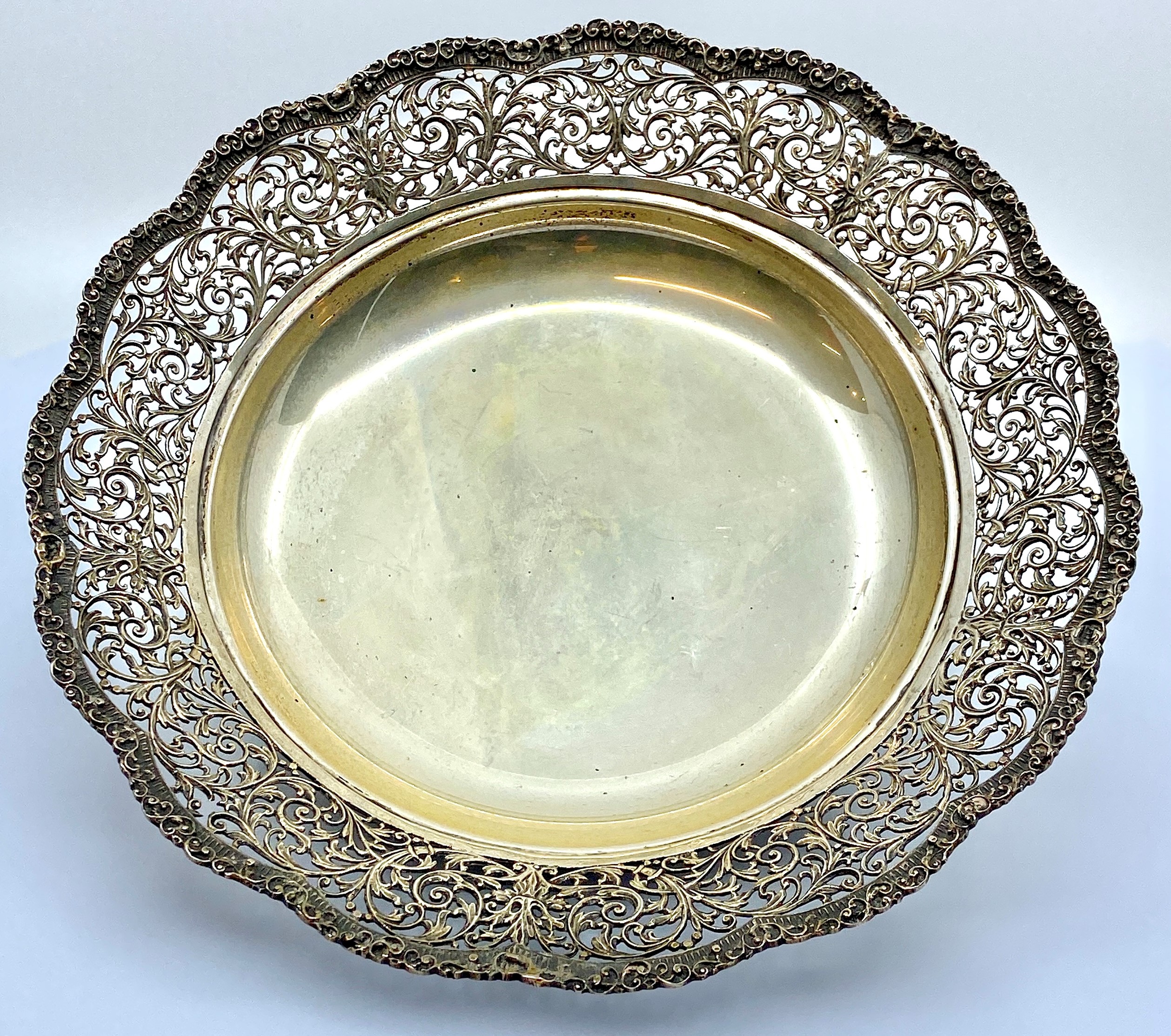 A white metal circular fruit dish with a pierced foliate scrolled border, standing on three