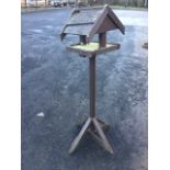 A wood birdtable with pitched roof, raised on square column stand with four angled legs.