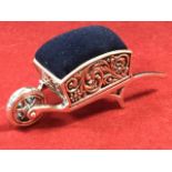 A silver pin cushion modelled as a wheelbarrow with foliate scroll decorated sides and spoked