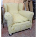 An early 20th Century wing back armchair upholstered in pale gold fabric, set on squat front bun