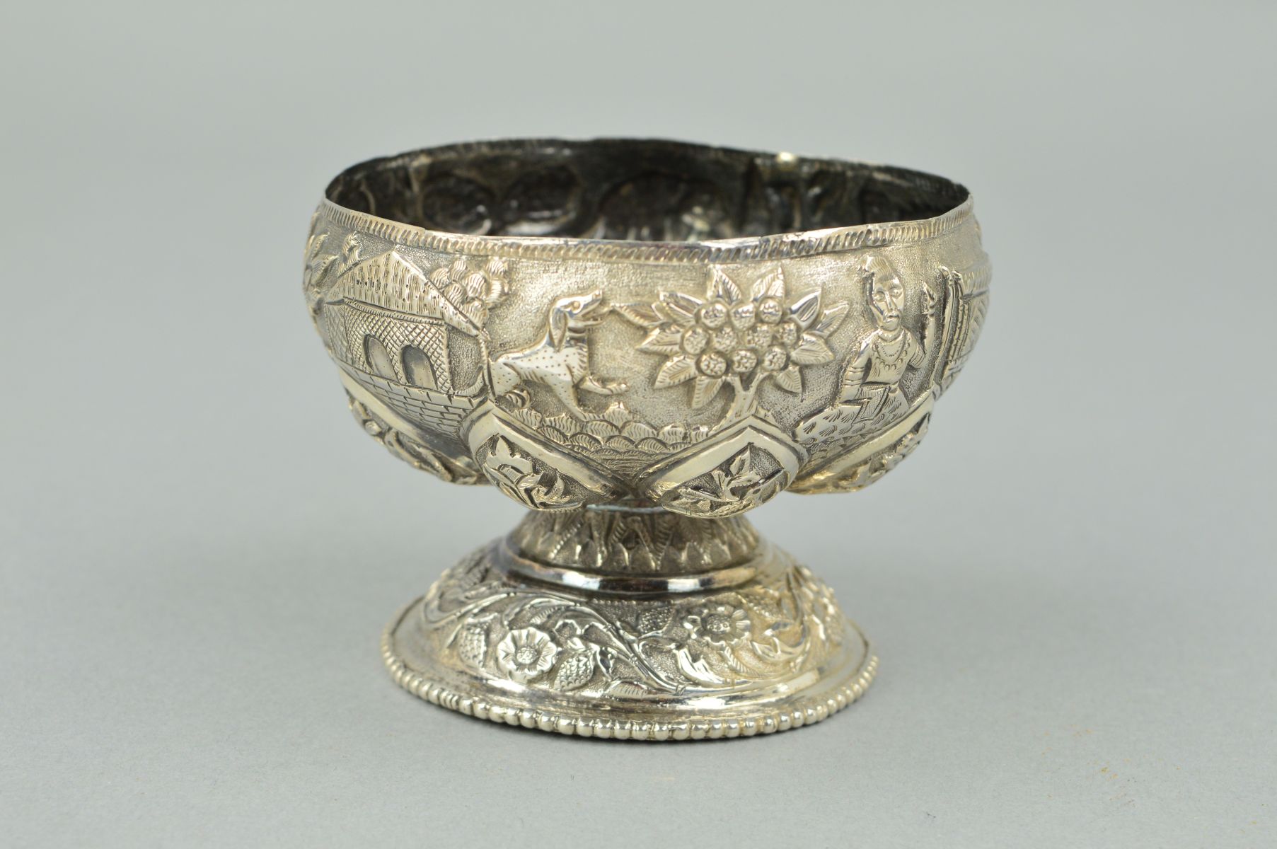 AN INDIAN WHITE METAL PEDESTAL BOWL, embossed decoration of animals, figures, foliage and buildings,