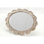 A WHITE METAL MIRROR OF OVAL FORM, the frame of wavy outline embossed with foliate scrolls, on a
